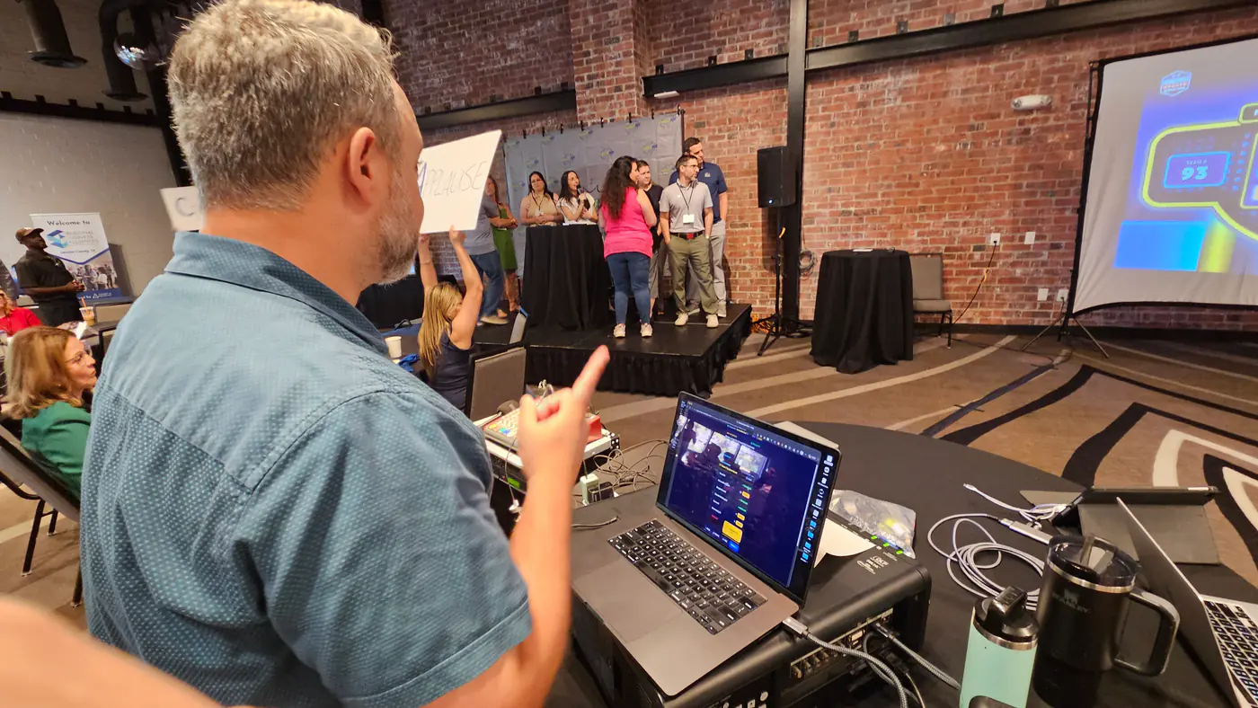 The host’s controls open on a laptop at the back of the room, the teams on stage, and the game board on the venue screen at the right.